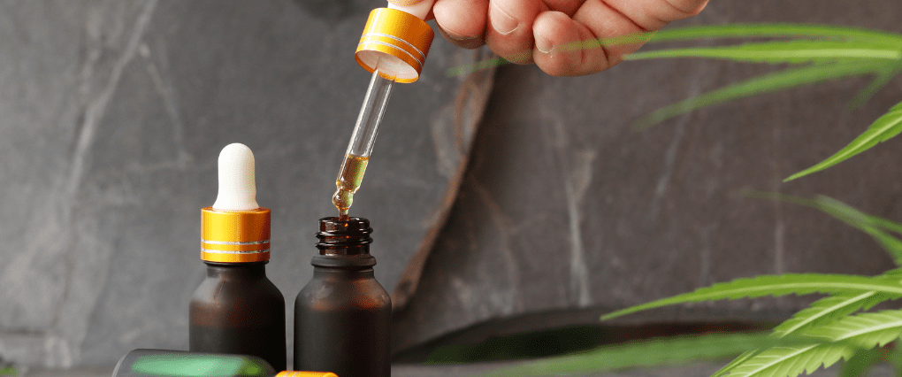 Hand opening CBD oil dropper bottle next to two CBD oil bottles on grey stone marble background