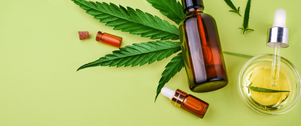 Flatlay image with bottle of CBD oil on top of hemp plant with pot of oil and CBD capsules against green background