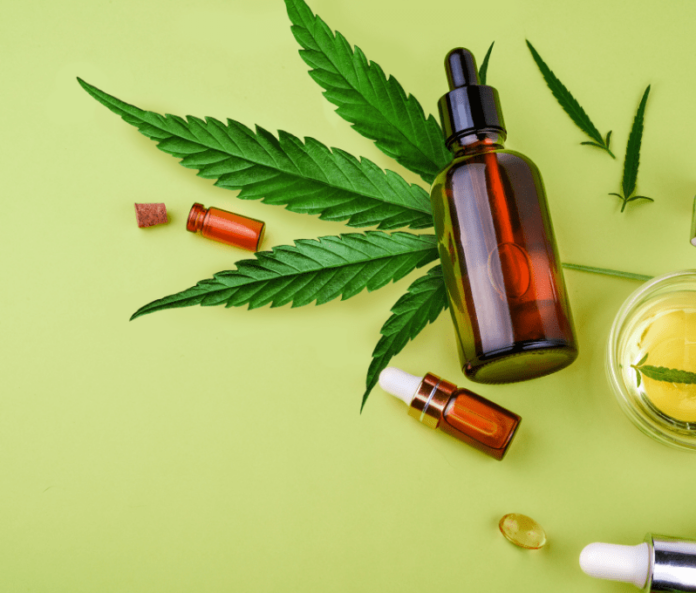 Flatlay image with bottle of CBD oil on top of hemp plant with pot of oil and CBD capsules against green background