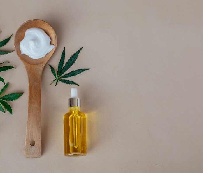 Flatlay image of moisturiser on a wooden spoon and CBD oil dropper bottle with hemp leaves