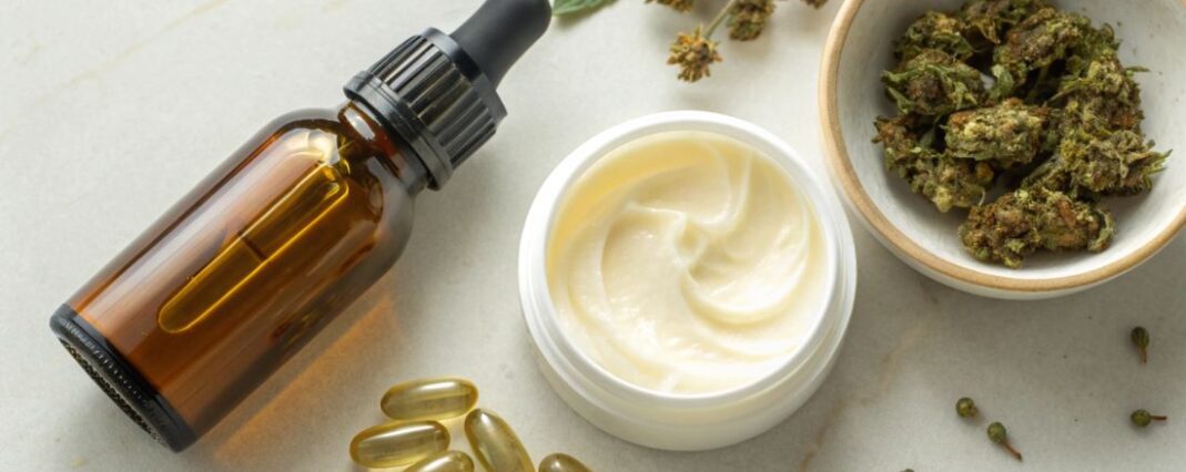 an oil dropper bottle, an open pot of cream, a bowl of cbd flower buds and some cbd capsules on a white surface
