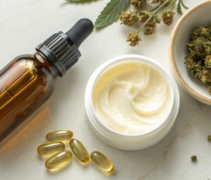 an oil dropper bottle, an open pot of cream, a bowl of cbd flower buds and some cbd capsules on a white surface