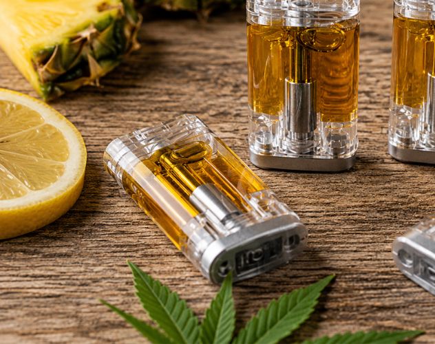 cbd vape pod surrounded by slices of lemon and pineapple and hemp leaves on a wooden surface